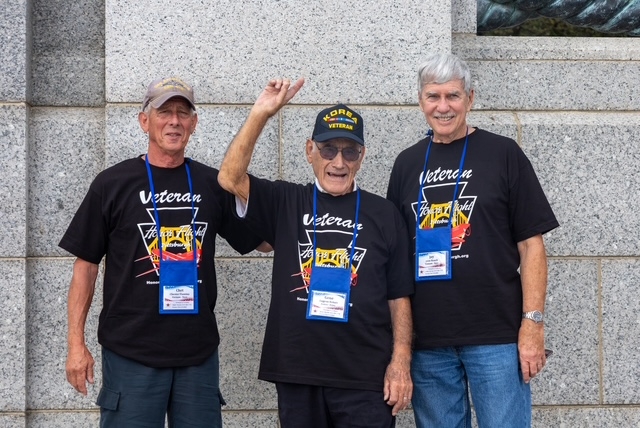 Three Veterans standing in front of an unidentfied memorial wall. One Veteran points overhead.
